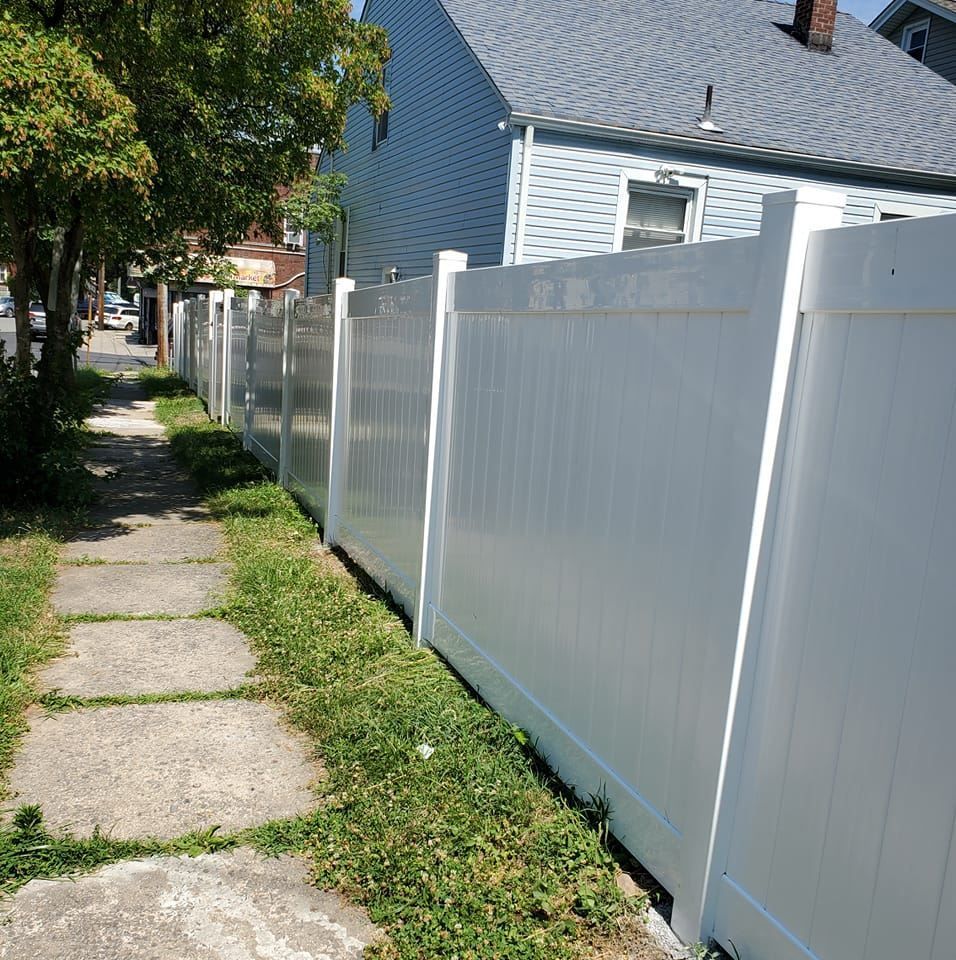 A white vinyl fence runs along a cracked concrete sidewalk next to a house on a sunny day.