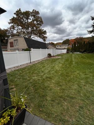 A grassy backyard featuring a white privacy fence, a stone border, and a large tree under a cloudy sky.