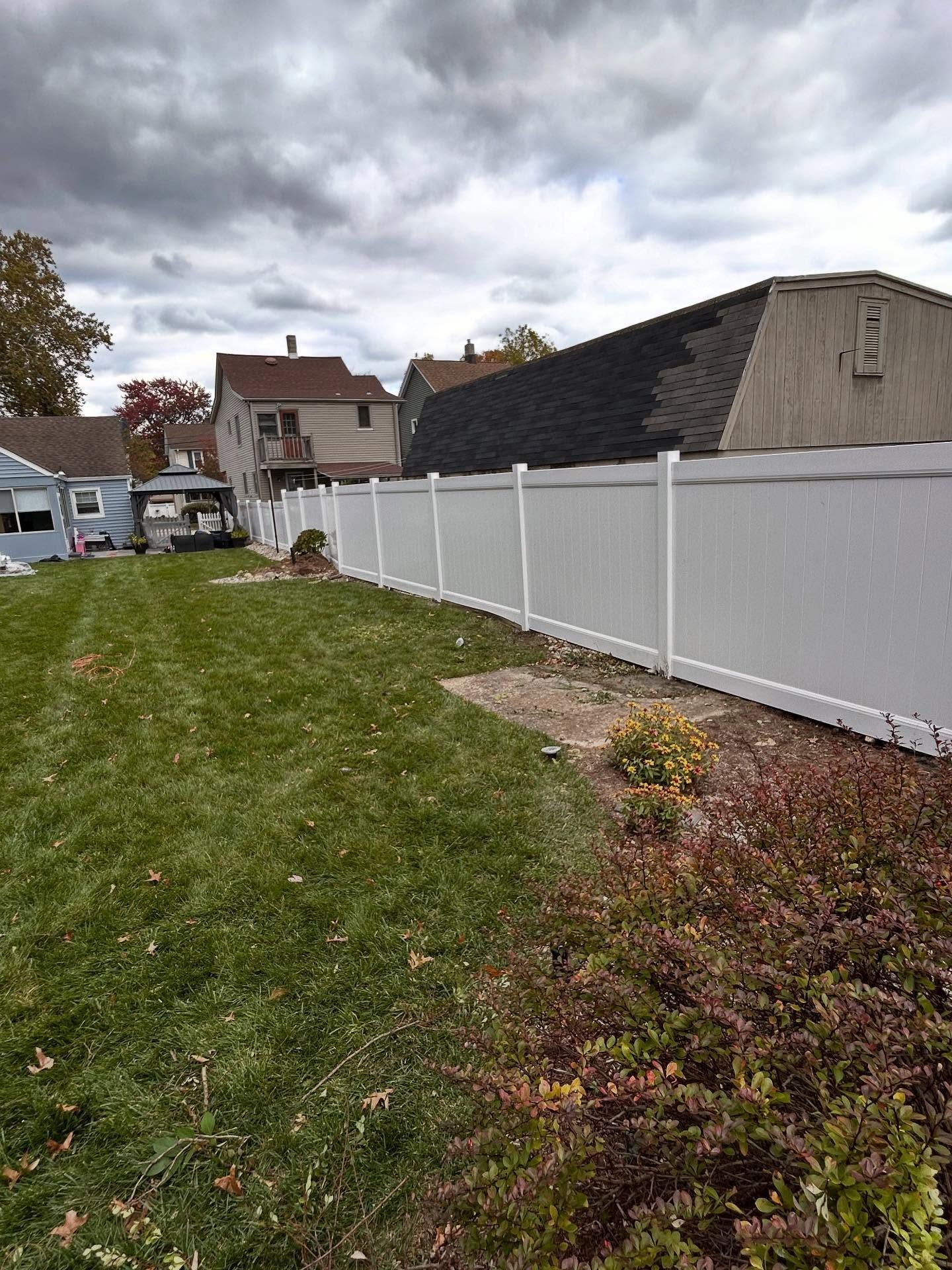 A white vinyl fence runs along a grassy yard with a beige building and houses in the background under a cloudy sky.