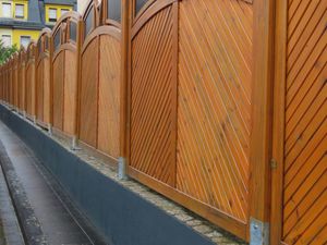 A series of wooden privacy fence panels with diagonal slats and arched tops, mounted on a low grey concrete base wall.