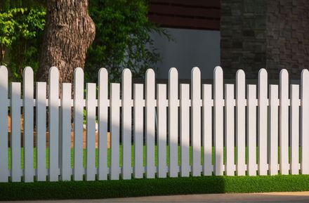 A clean, white picket fence stands in front of a green lawn and a tree, with light and shadow cast across the slats.