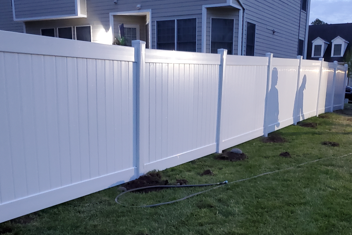A long, white vinyl privacy fence stands in a grassy backyard next to the side of a house.