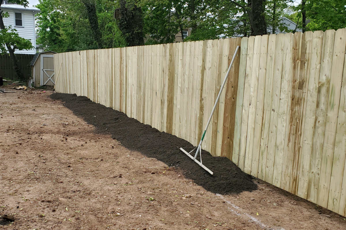 A rake rests against a new light-colored wooden privacy fence, next to a long pile of dark soil in a dirt yard.