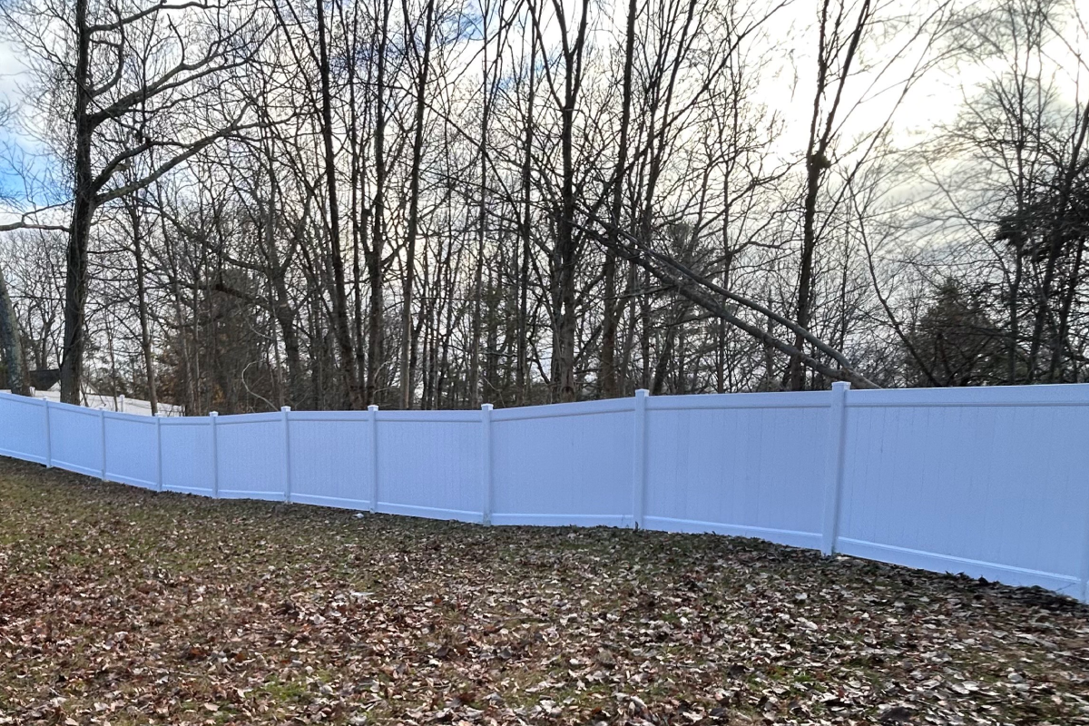 A white vinyl privacy fence runs across a yard covered in fallen brown leaves, with a line of bare trees in the background.