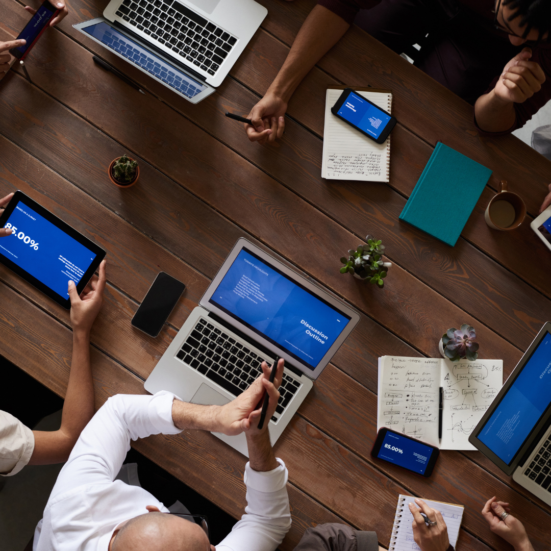 People at a table with laptops and tablets during a meeting, some raising hands.