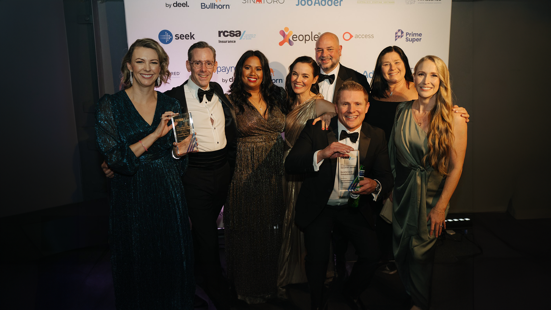 Group of people in formal wear at an awards ceremony, holding trophies in front of a backdrop with logos.