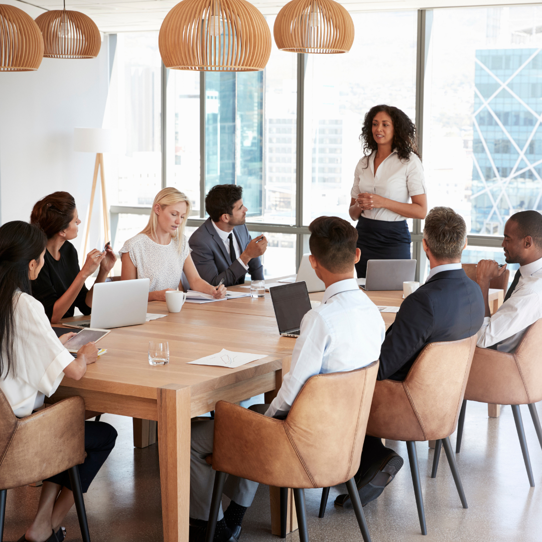 Business meeting in a bright room. A woman addresses seated colleagues around a large table; laptops, windows.