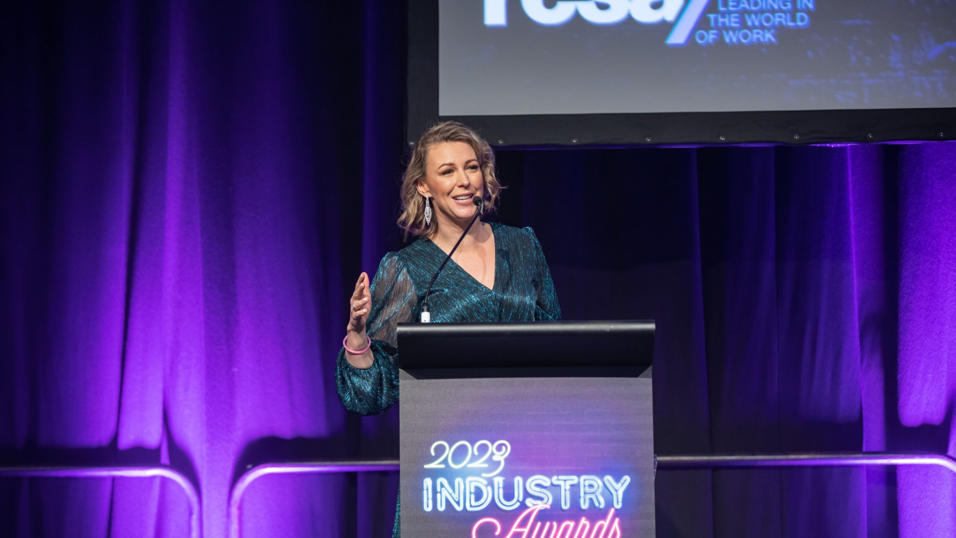 Woman speaking at a podium, at an awards event with a purple backdrop.