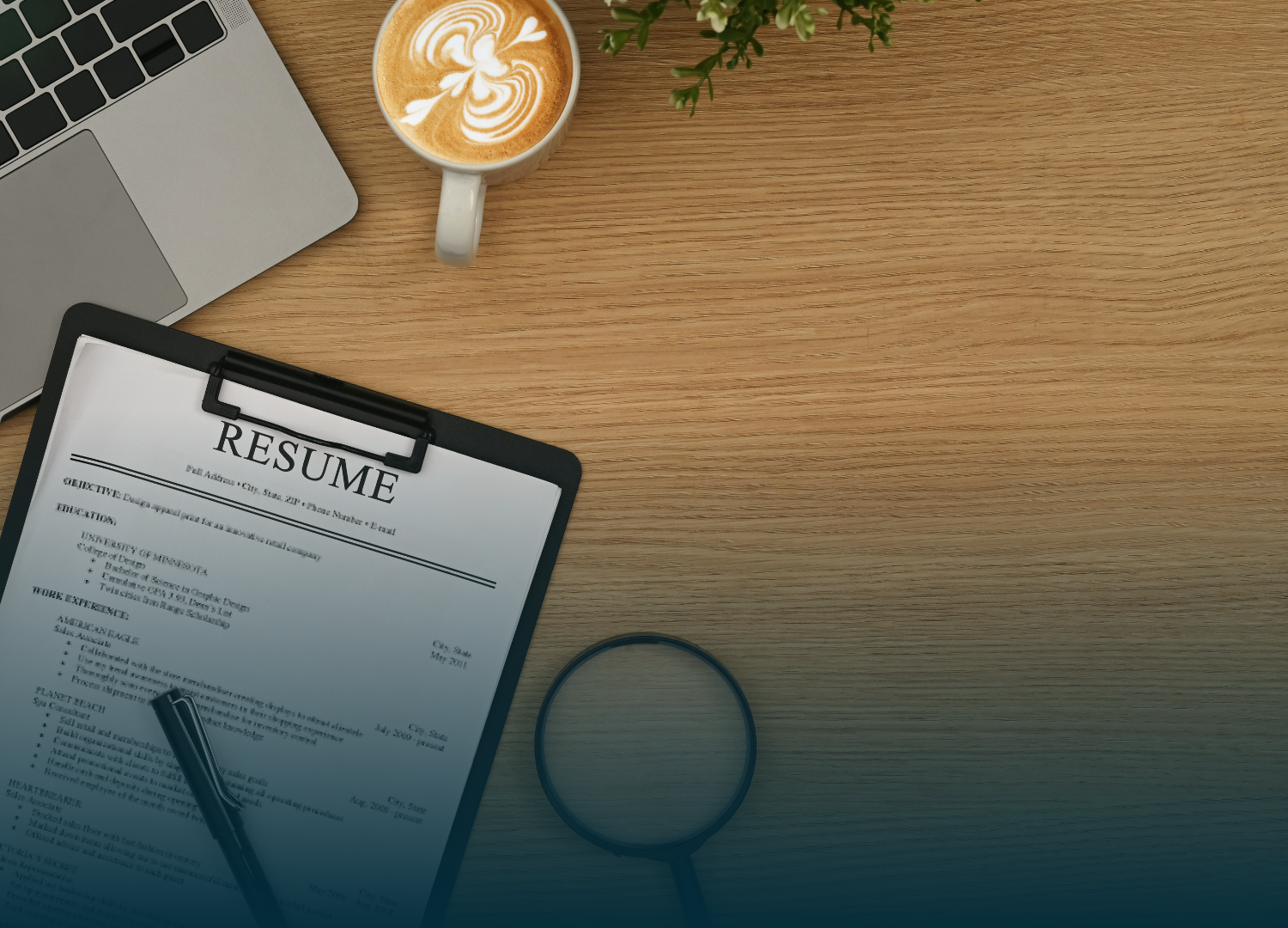 Laptop, resume on clipboard, coffee cup, pen, magnifying glass, and small plant on a wooden desk.