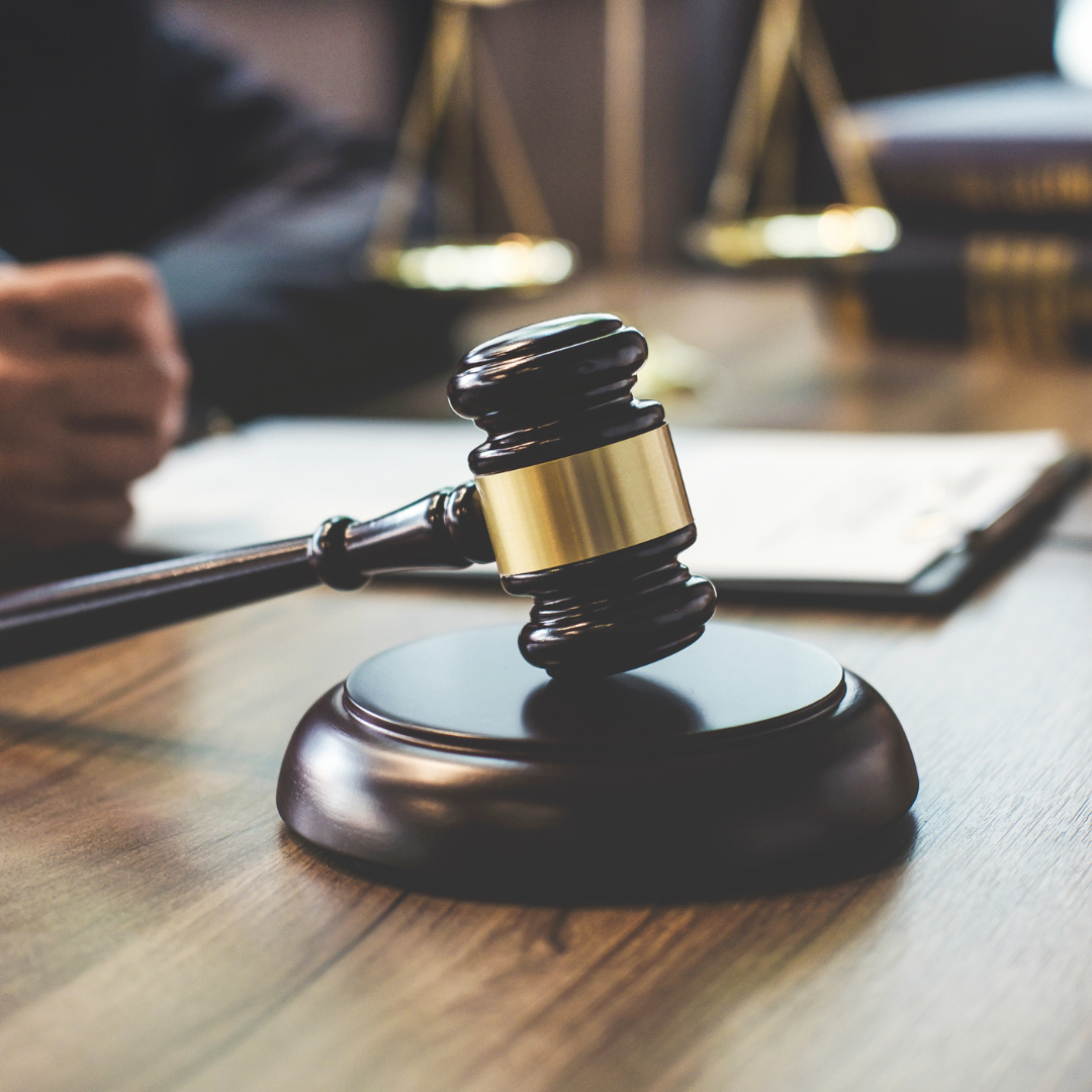 Gavel on wooden block; scales and documents in background, legal setting.