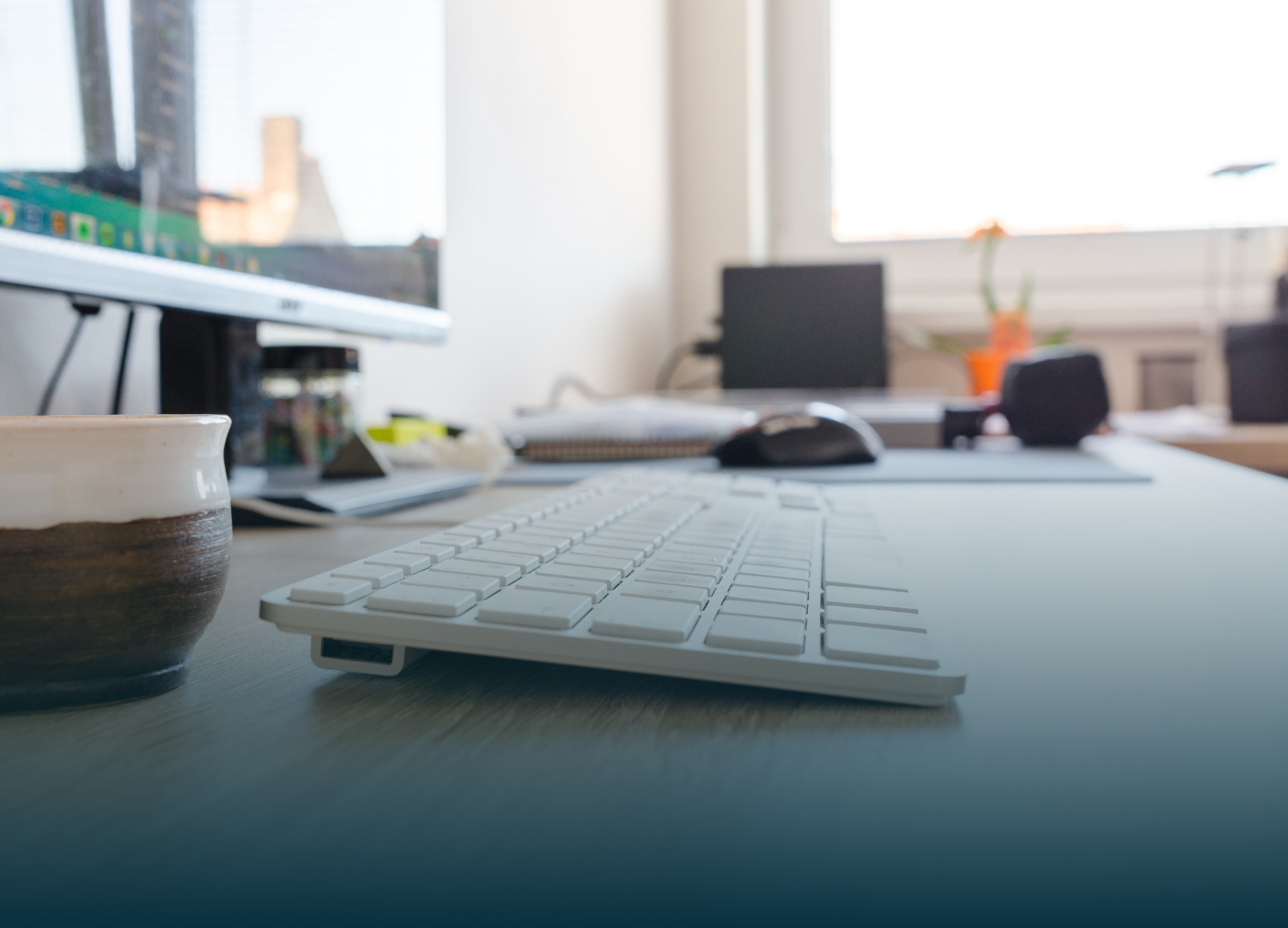 White keyboard on a desk, with a mug, monitor, and laptop in the background near a window.