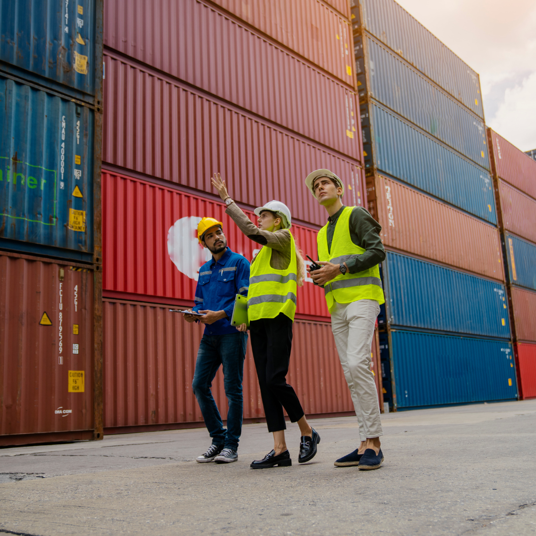 Three people in safety vests at a shipping port inspect stacked containers; one points upwards.