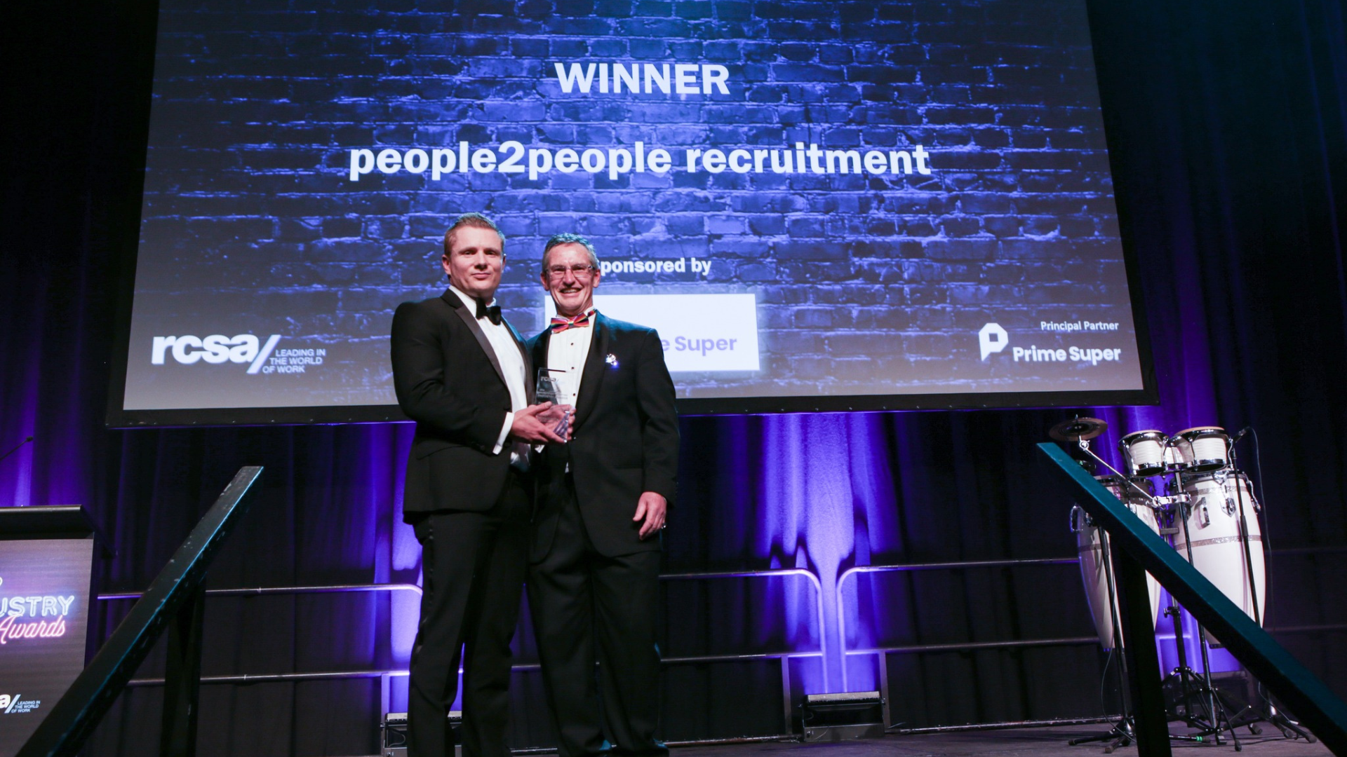 Two men on stage holding an award, with a screen behind them that says 