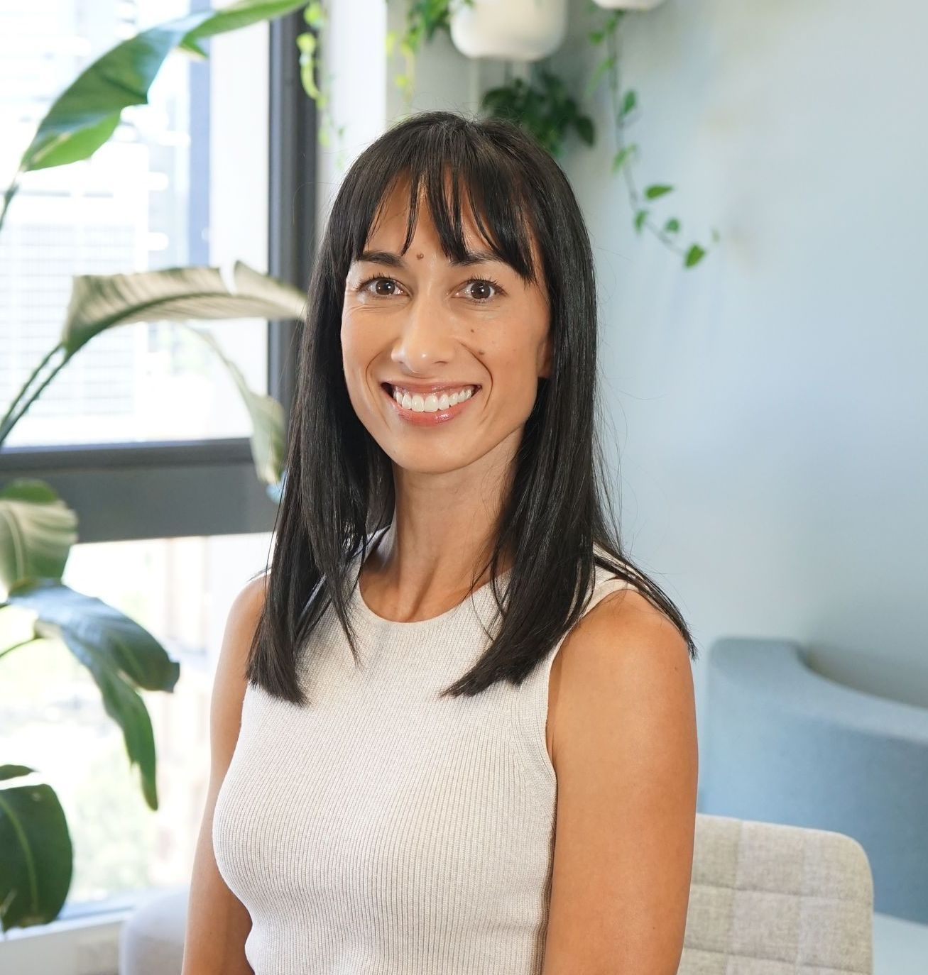 Woman with black hair smiles, seated near a window with plants, wearing a white textured sleeveless top.