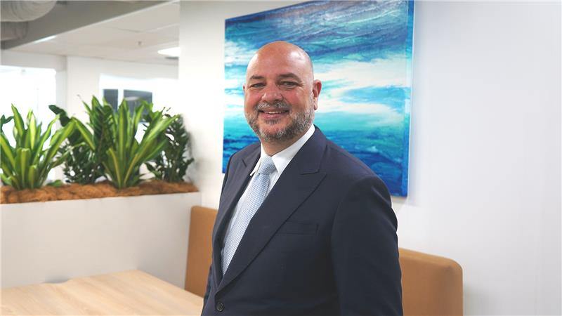 A smiling person in a dark suit and light tie stands in an office with green plants and blue abstract art in the background.