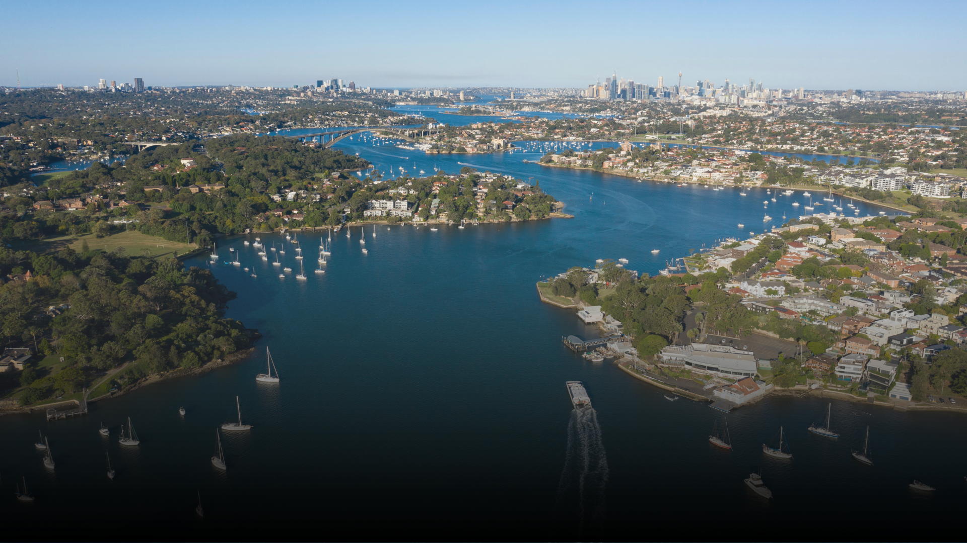 Aerial view of a harbor with numerous sailboats, houses, and city skyline in the background. Blue water and a sunny sky.