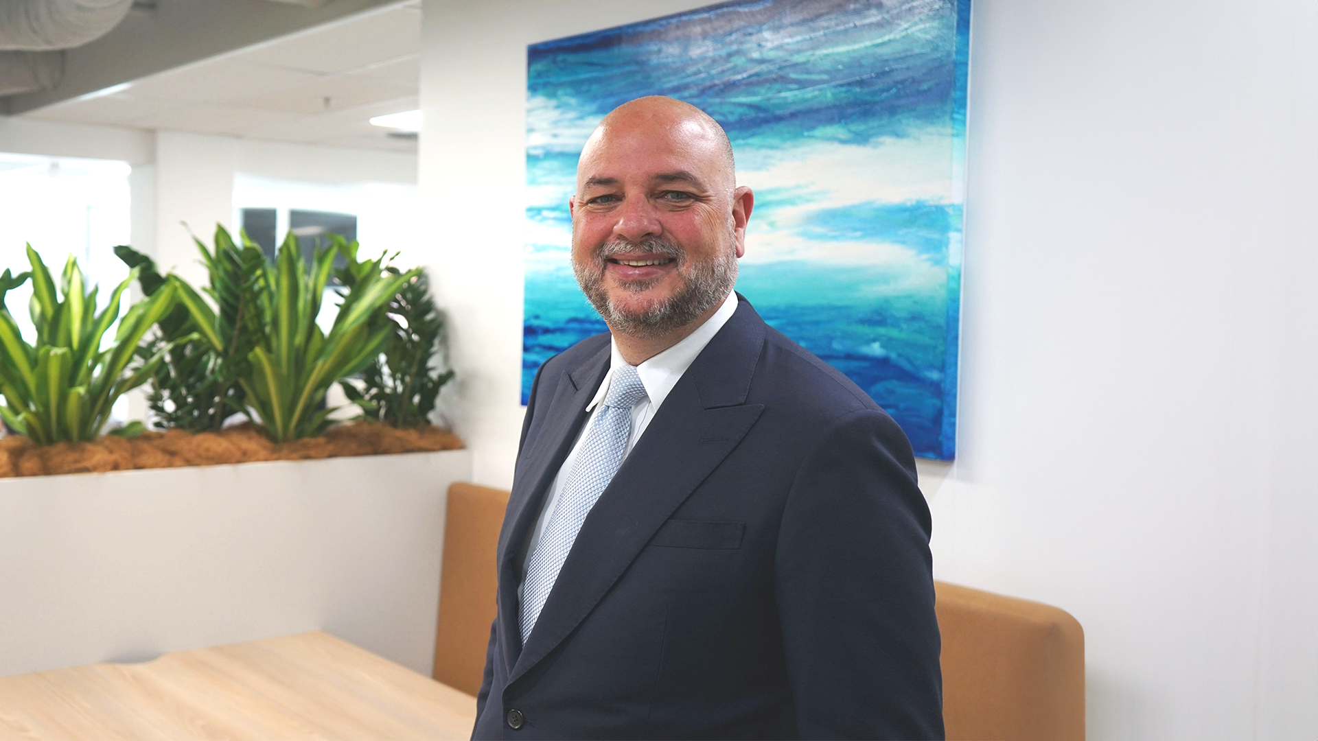 A smiling professional in a suit and tie standing in a modern office with greenery and blue abstract art in the background.
