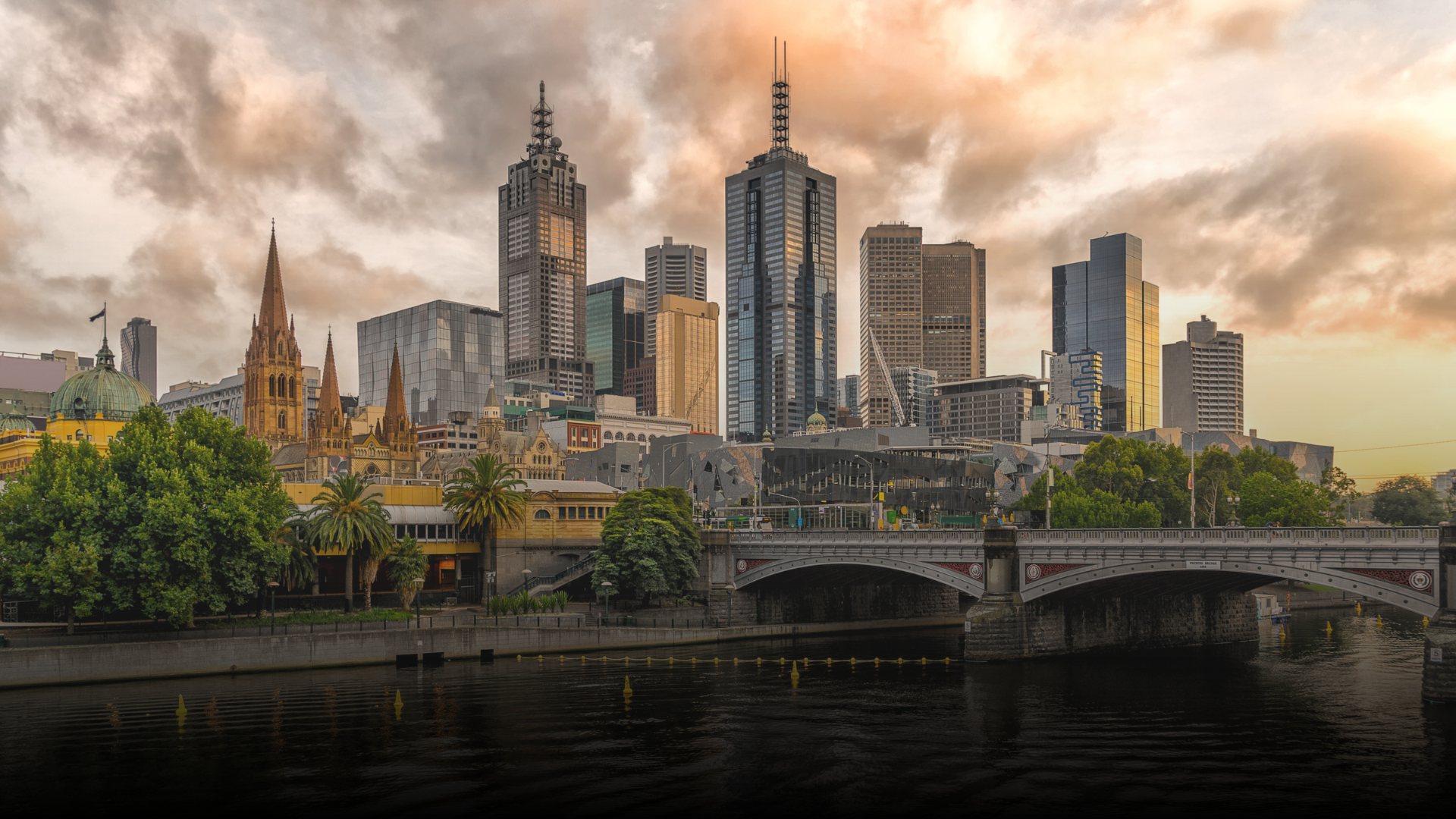 Melbourne skyline at sunset, with buildings, bridge, river, and cloudy sky.