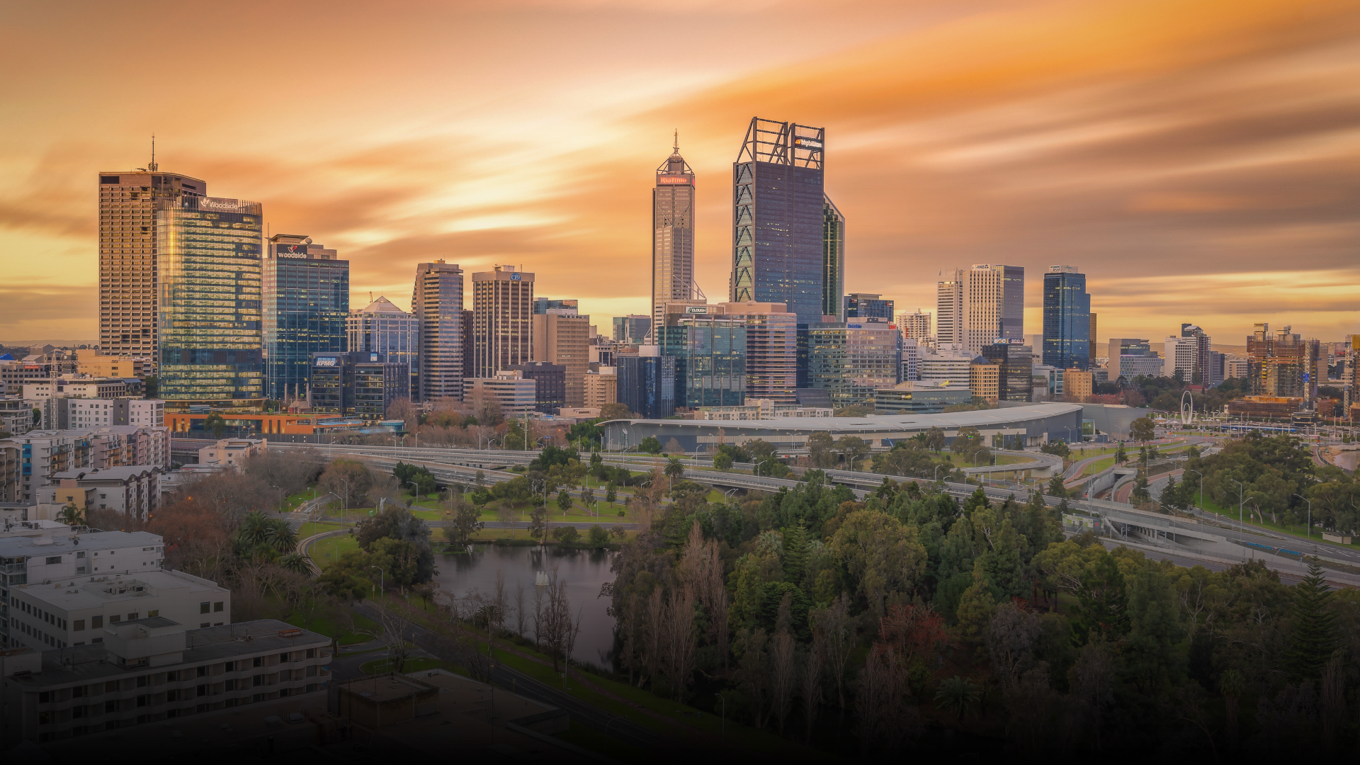 Perth, Australia skyline at sunset, featuring tall buildings and a river. Orange sky.