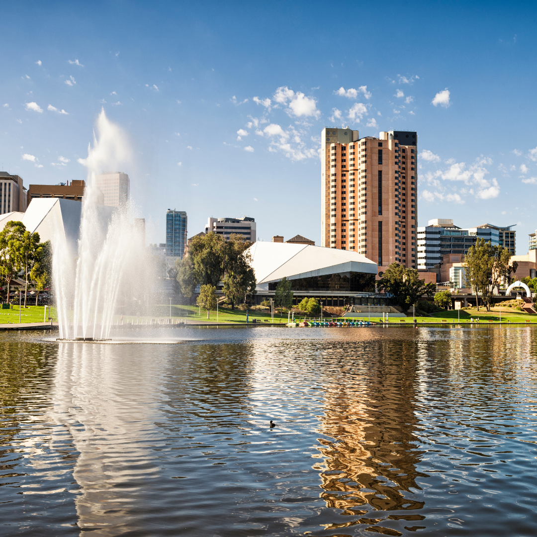 Fountain spraying water in a lake with city buildings in the background under a blue sky.