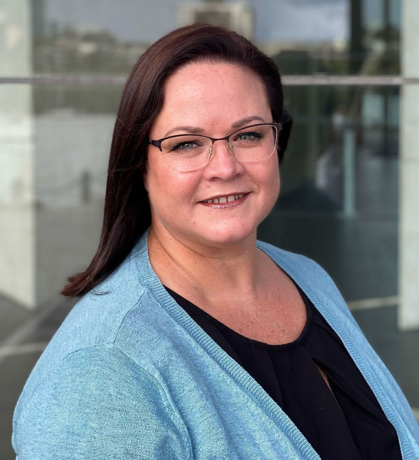 Woman with glasses smiles; wearing blue sweater and black shirt, in front of glass building.