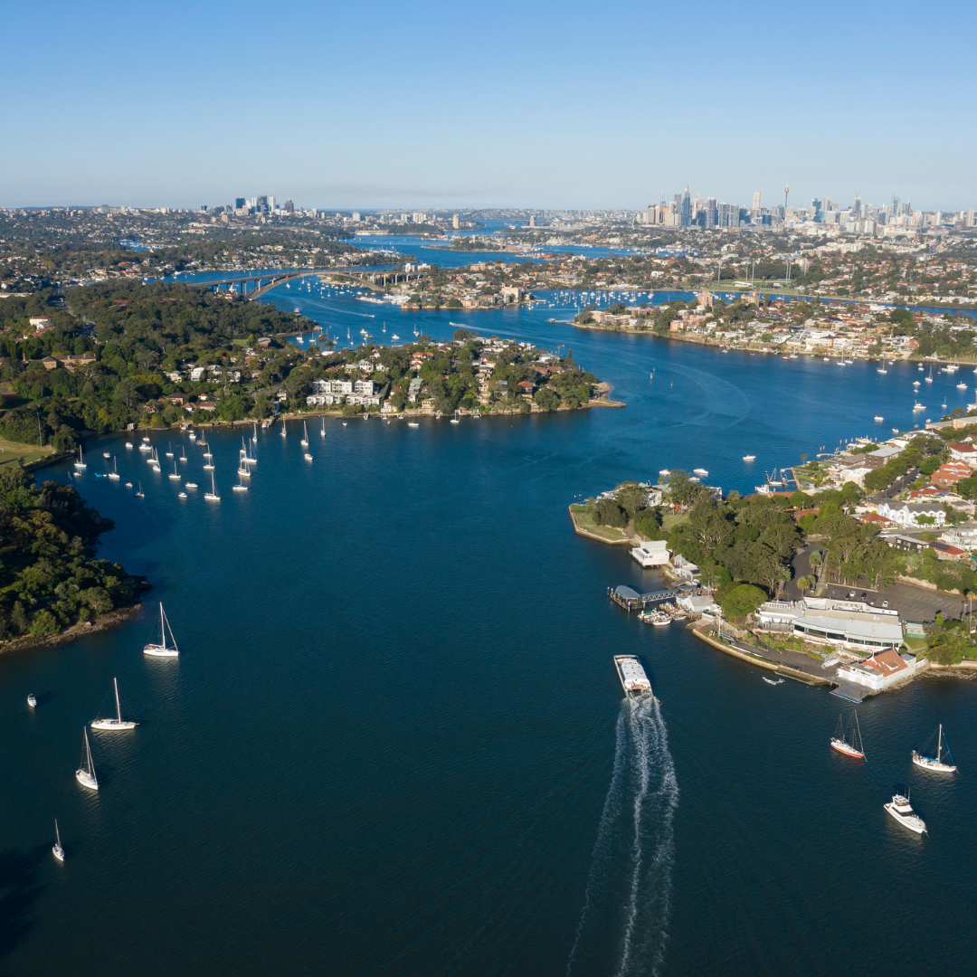 Aerial view of Sydney Harbour with boats, lush green islands, and city skyline under a clear blue sky.