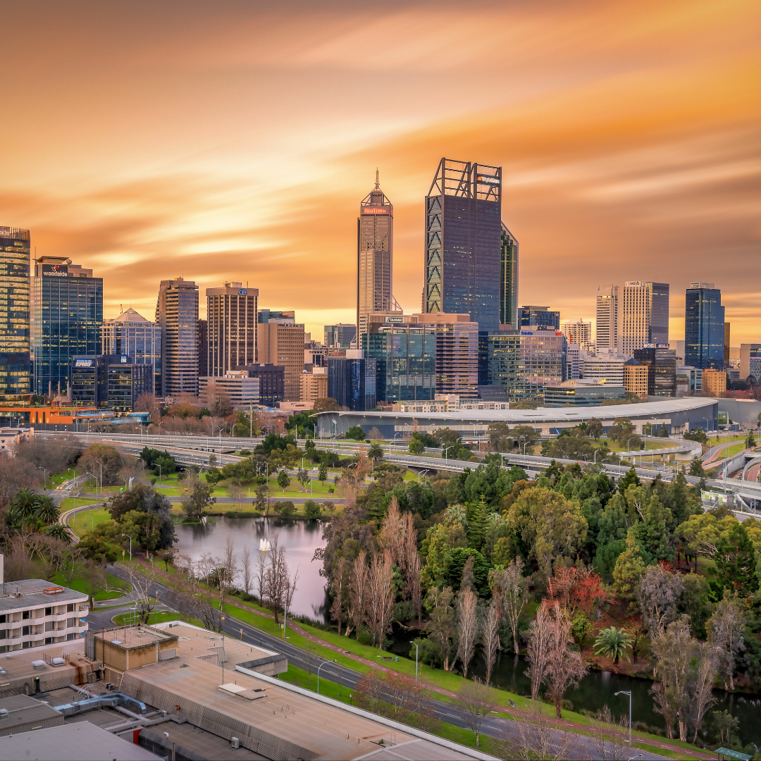 Perth, Australia skyline at sunset, featuring city buildings, a lake, and lush greenery under an orange sky.