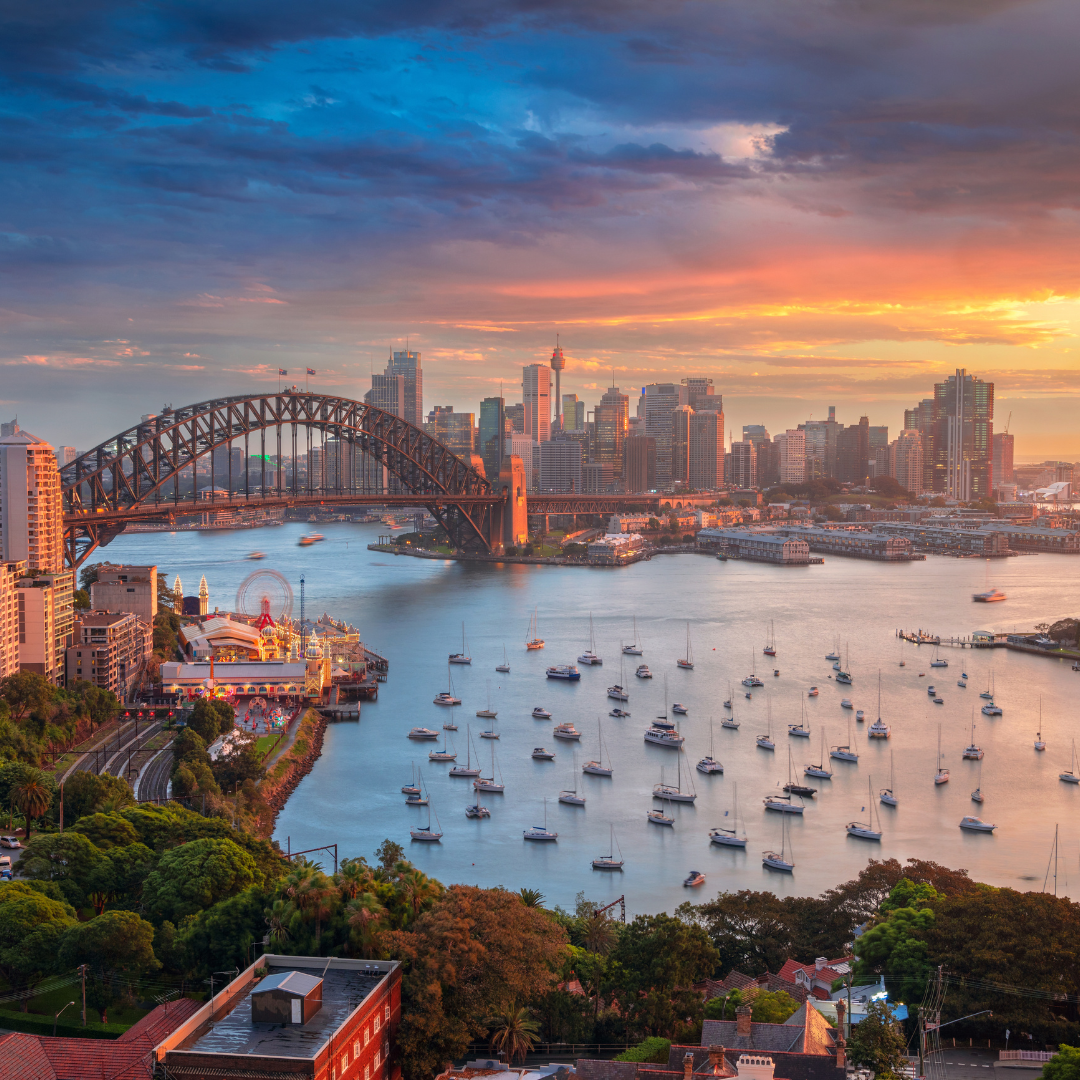 Sydney Harbour at sunset with boats, bridge, and city skyline.