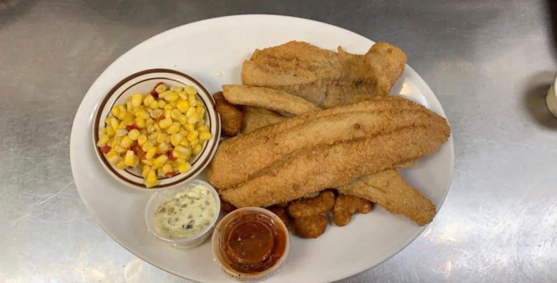 Plate of fried fish fillets with corn, coleslaw, and dipping sauces on a metal table