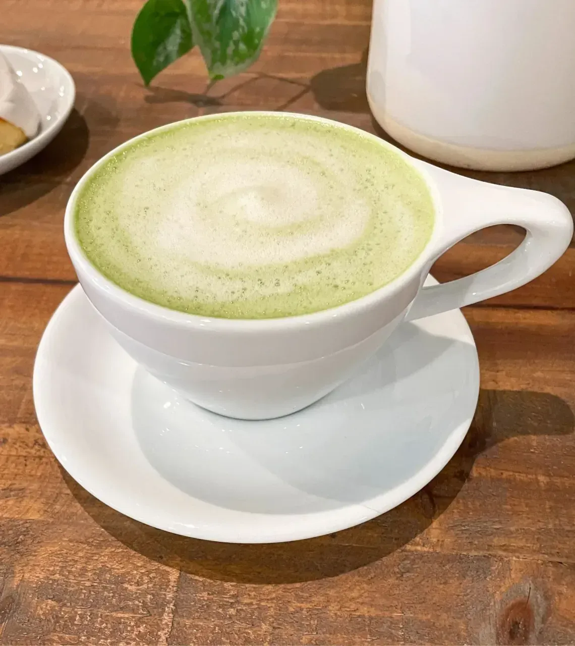 White cup of green matcha latte on a saucer atop a wooden table