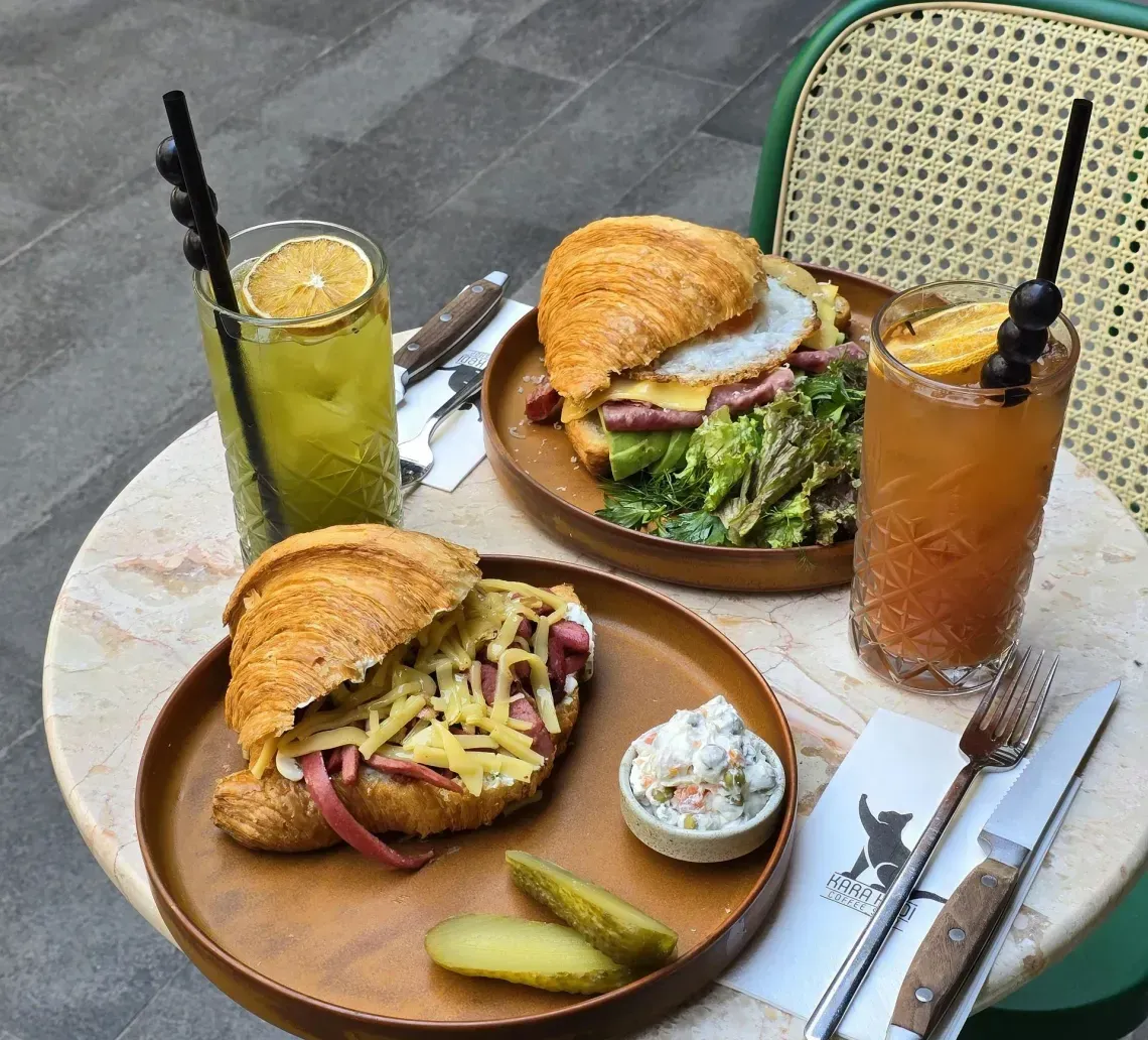 Two sandwiches with pickles and two iced drinks on a small outdoor café table