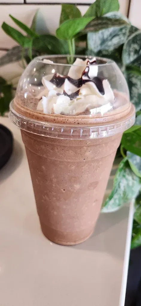 Chocolate smoothie topped with whipped cream and chocolate drizzle in a clear plastic cup, on a table with plants behind it