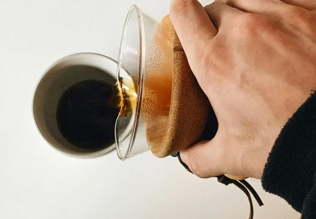 Hand pouring coffee from a cup into a glass mug with a brown sleeve