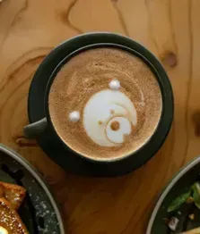 Top-down view of a latte in a green mug on a wooden table, with food plates partly visible nearby