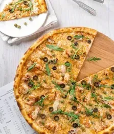 Overhead view of a vegetable pizza on a wooden board with a slice removed, beside a plate and utensils