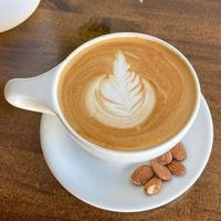White cup of latte with leaf latte art on a saucer, beside almonds on a wooden table.