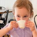 Child drinking from a small white cup at a table, wearing a lavender shirt.
