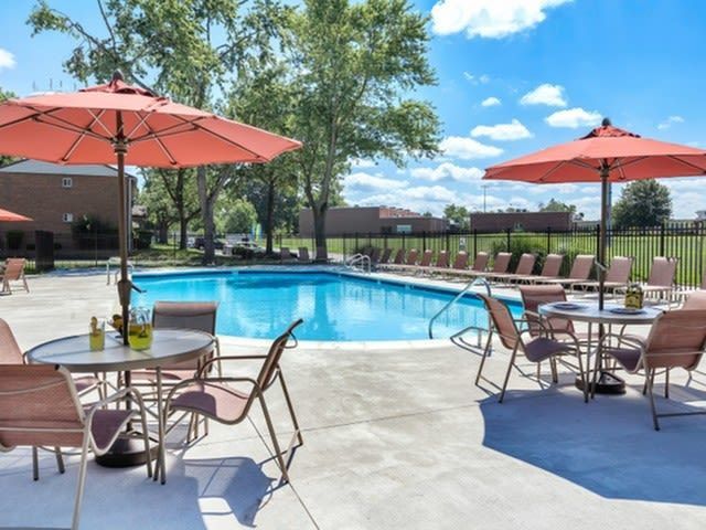Outdoor apartment community pool area with lounge chairs, tables, and umbrellas under blue skies.