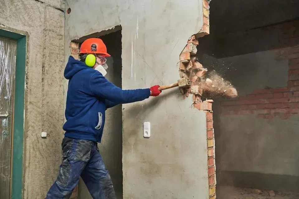 Ein Arbeiter in Schutzhelm und Schutzkleidung reißt mit einem Vorschlaghammer eine Ziegelmauer ein.