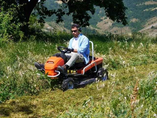 Una mietitrebbia gialla al lavoro in un campo di grano, accanto ad alberi verdi e a un edificio.