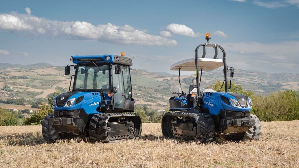Due trattori blu in un campo arido con colline e nuvole sullo sfondo.