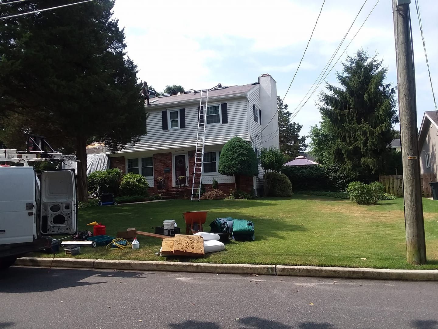 Two-story white house with ladder on roof, yard with construction materials, trees, sunny day.