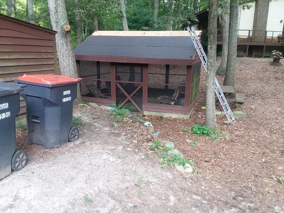 Chicken coop with dark roof and brown frame, near trash bins and trees.
