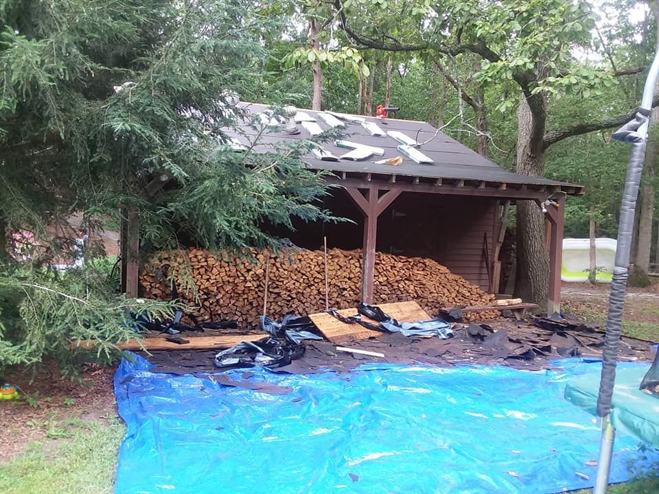 Wooden shed filled with firewood, roof damage, blue tarp on ground. Forest setting.