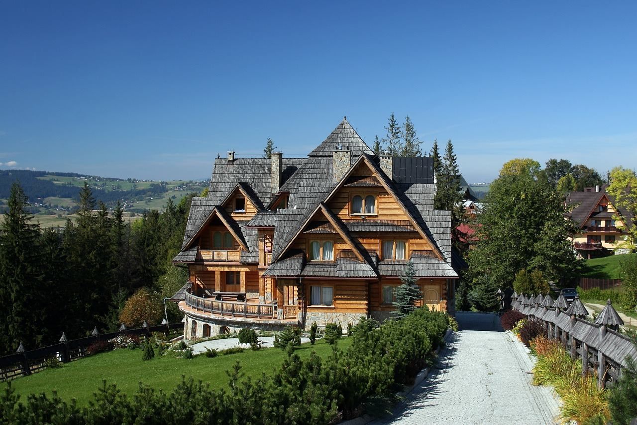 Wooden house with a steeply pitched shingled roof, set amidst green trees with mountains in the background under a blue sky.