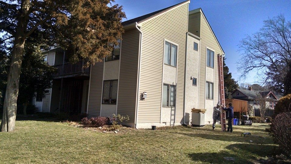 Two-story tan and white building with a grassy lawn, a tree, and two people near a ladder.