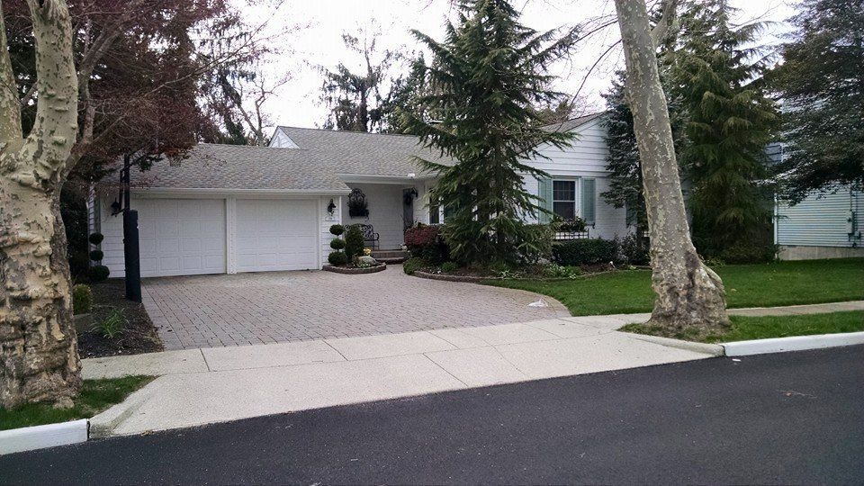 White house with two-car garage, brick driveway, and front lawn. Trees line the side of the home and road.