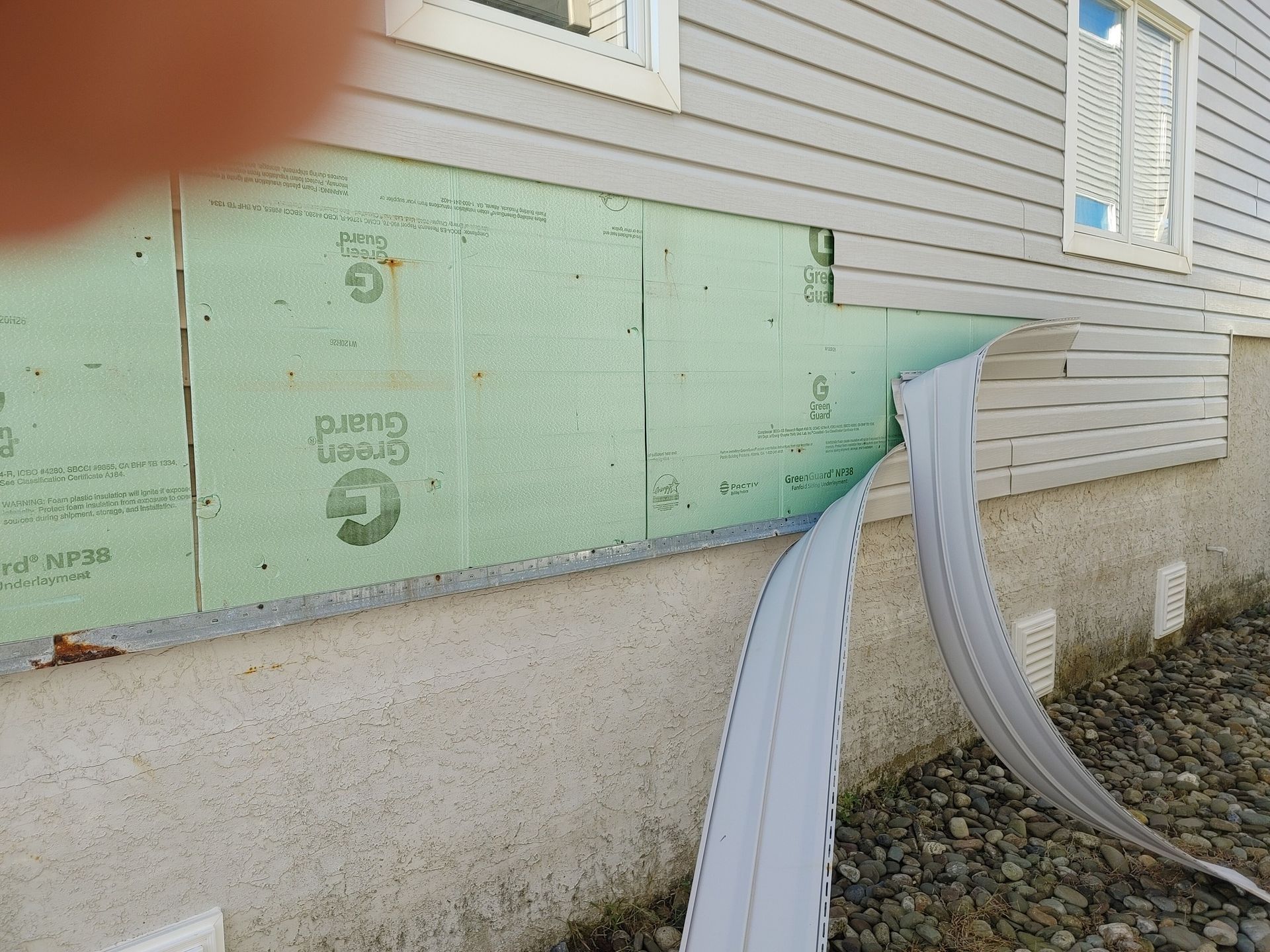 Damaged house siding with exposed green insulation panels, detached gutter, concrete foundation.
