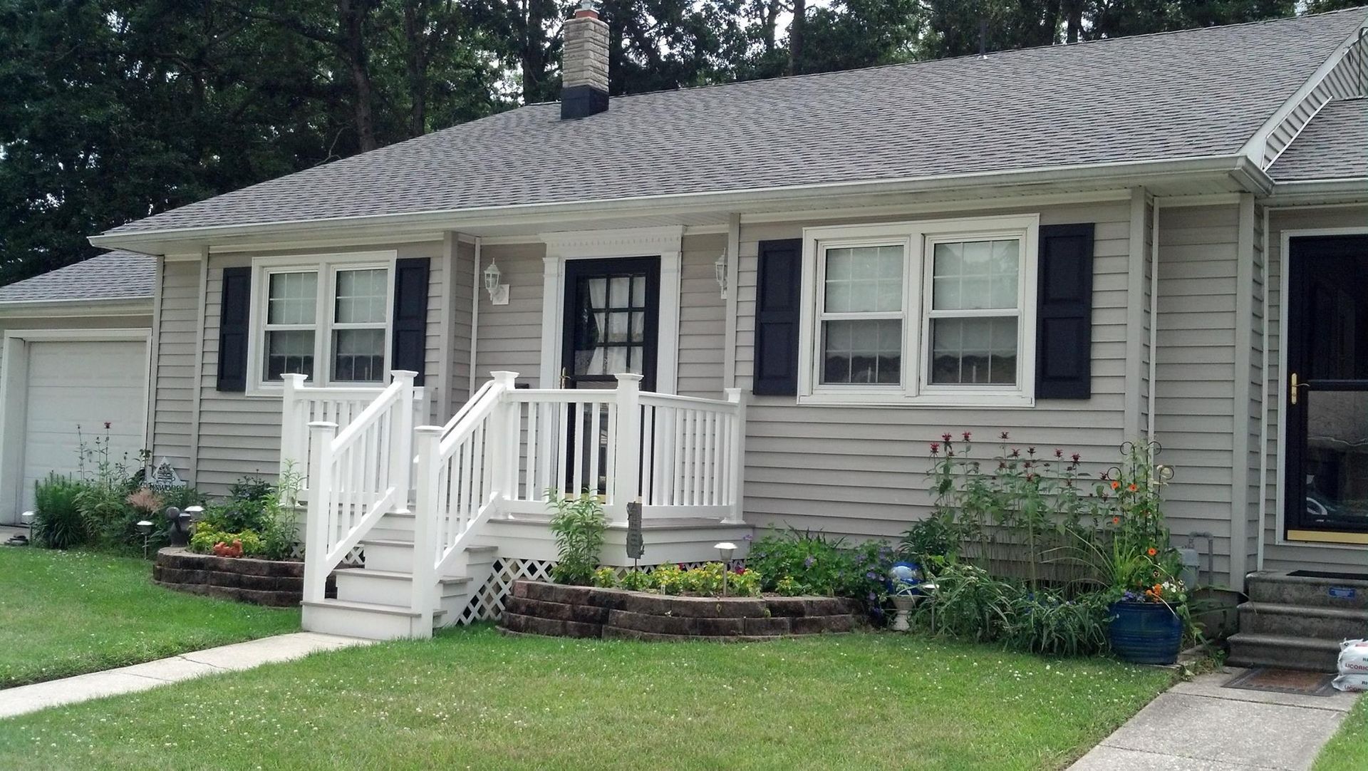 Gray house with small porch, white railing, black shutters, and small garden in front.