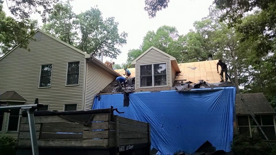 Roofers working on a house, covering damaged area with blue tarp. Trees in background, overcast day.